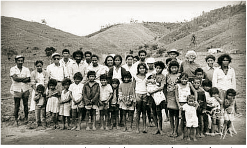 Fotografia em preto e branco do povo indígena Krenak reunido, com homens, mulheres e crianças posando a céu aberto em frente a colinas.