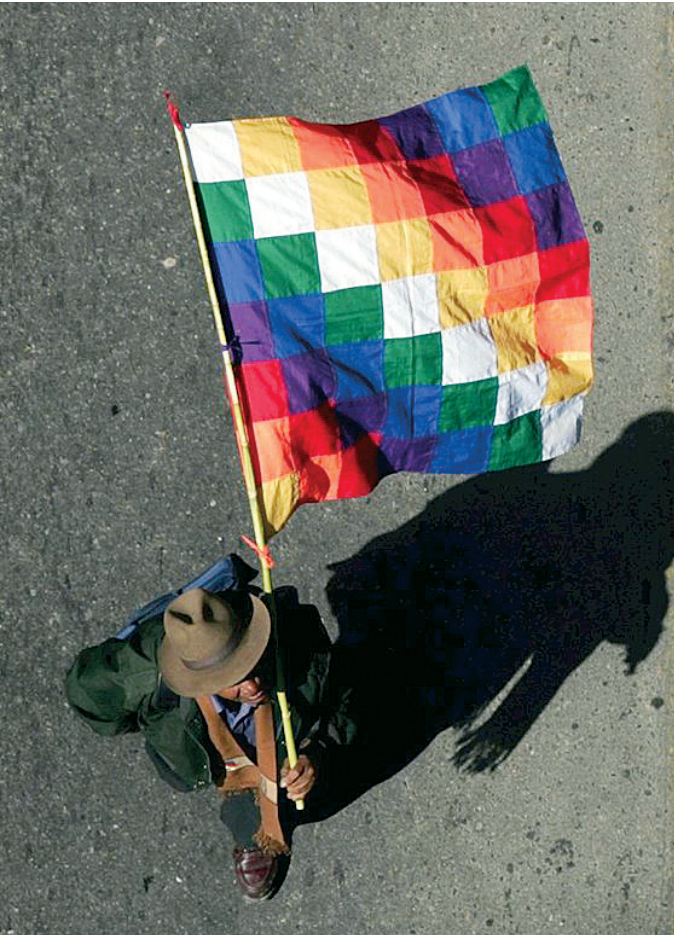 Fotografia em vista aérea de uma pessoa caminhando com chapéu sobre o asfalto, projetando uma longa sombra à direita e segurando uma haste com a bandeira Wiphala estendida ao vento, formada por um padrão xadrez diagonal multicolorido.