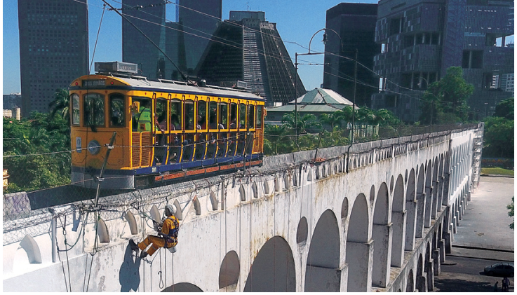Bonde trafegando sobre os Arcos da Lapa, no Rio de Janeiro, com edifícios contemporâneos e a Catedral Metropolitana ao fundo