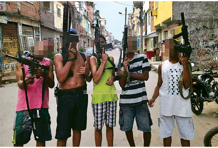 Grupo de cinco jovens em pé em uma rua de favela portando fuzis e armas de fogo, com os rostos cobertos por efeito de pixelização.