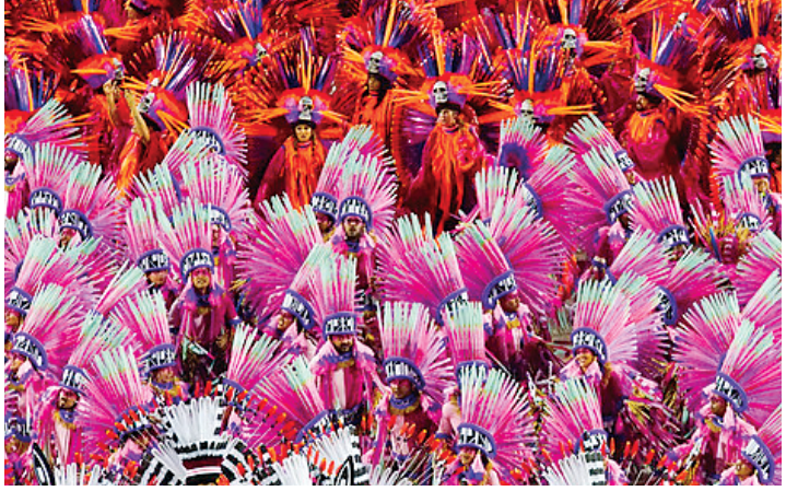 Integrantes de ala de escola de samba usando fantasias volumosas com penas e adereços nas cores rosa, roxo e vermelho durante desfile.