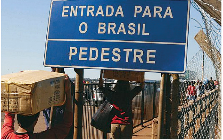 Pessoas carregando fardos e caixas sobre a cabeça atravessando passagem de pedestres sob uma grande placa azul com os dizeres: "ENTRADA PARA O BRASIL PEDESTRE".