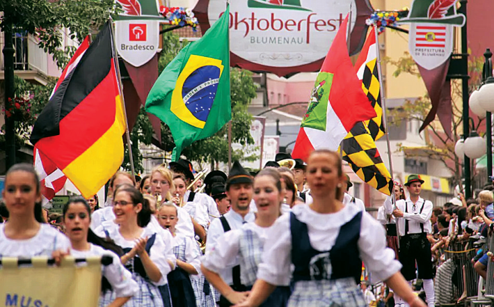 Desfile tradicional da festa Oktoberfest em Blumenau, com pessoas em trajes alemães típicos carregando as bandeiras da Alemanha, do Brasil e do município.