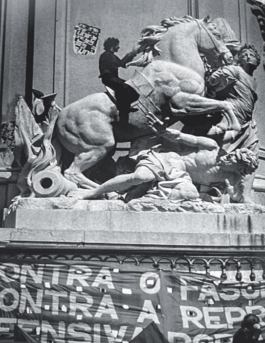 Manifestante montado sobre a escultura de um cavalo em um monumento público decorado com faixa de protesto.