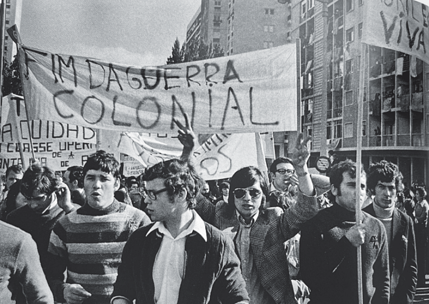 Manifestantes marchando nas ruas segurando uma grande faixa com os dizeres "FIM DA GUERRA COLONIAL".