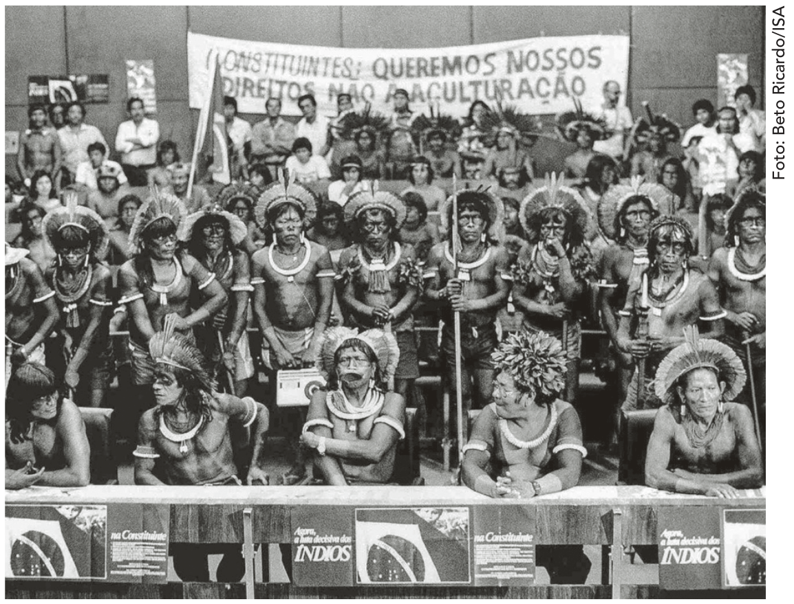 Fotografia em preto e branco de lideranças indígenas de várias etnias reunidas em auditório durante a Assembleia Constituinte de 1988. Ao fundo, uma faixa com os dizeres: "CONSTITUINTES: QUEREMOS NOSSOS DIREITOS NÃO A ACULTURAÇÃO". Na bancada frontal, cartazes com os dizeres "na Constituinte" e "Agora, a luta decisiva dos ÍNDIOS".