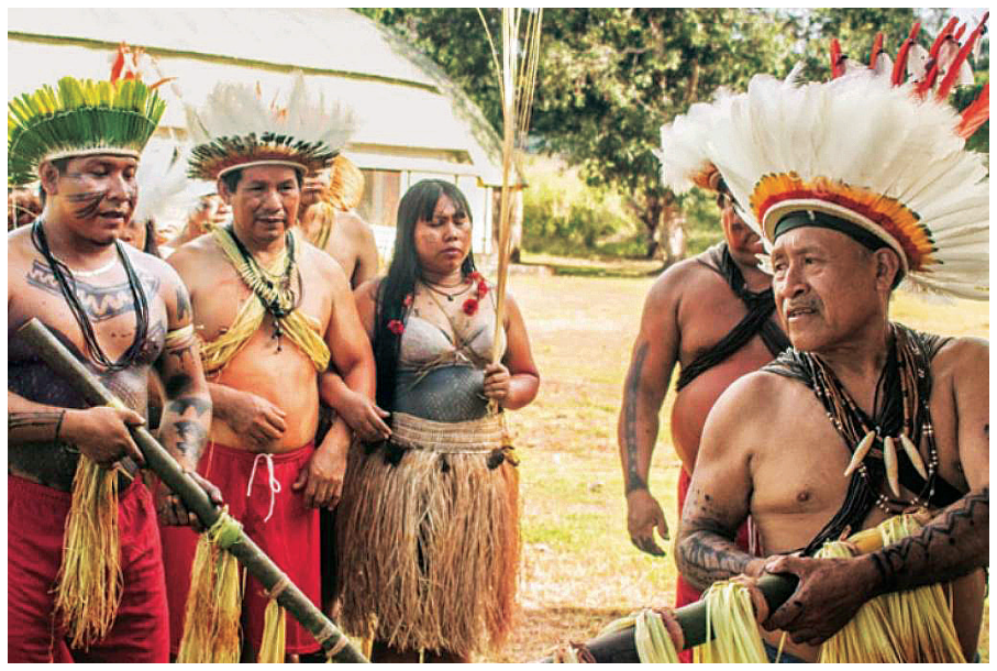 Indígenas do povo Zoró com adereços tradicionais, como cocares de penas, colares e pinturas corporais, segurando varas de bambu.