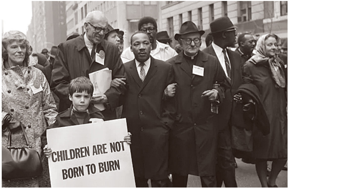 Martin Luther King Jr. e outros manifestantes marcham em protesto de braços dados; à frente, um menino segura um cartaz onde se lê "CHILDREN ARE NOT BORN TO BURN".