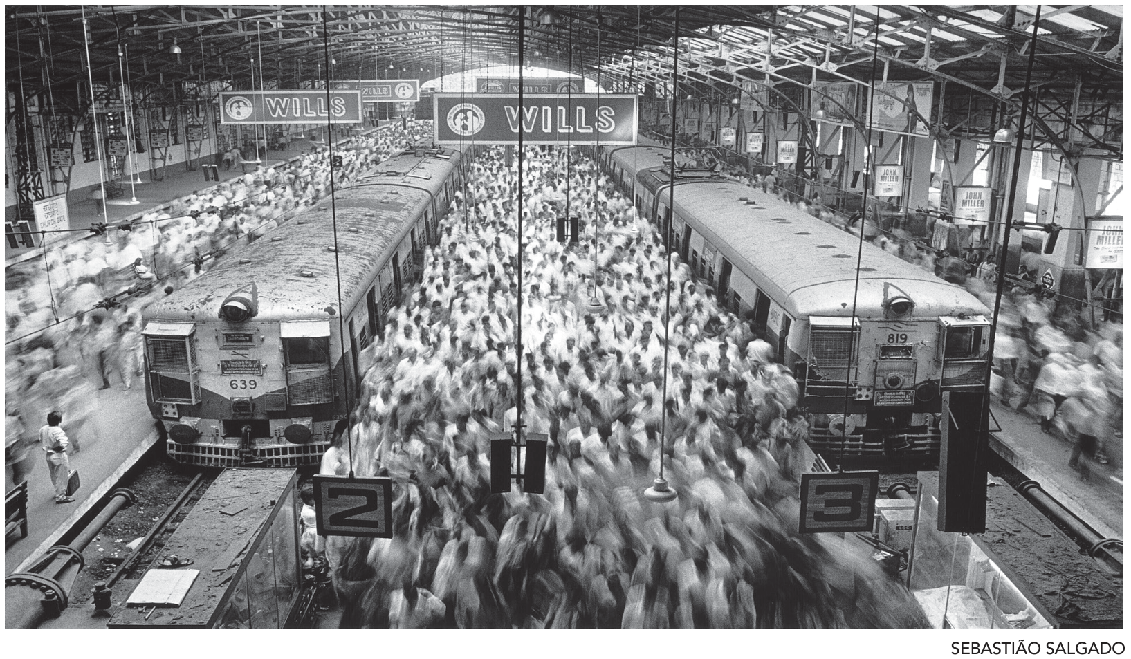 F1 Fotografia em preto e branco com vista superior da Estação de Trem Churchgate em Mumbai, mostrando uma grande multidão em movimento borrado entre trens estacionados nas plataformas cobertas por estrutura metálica.