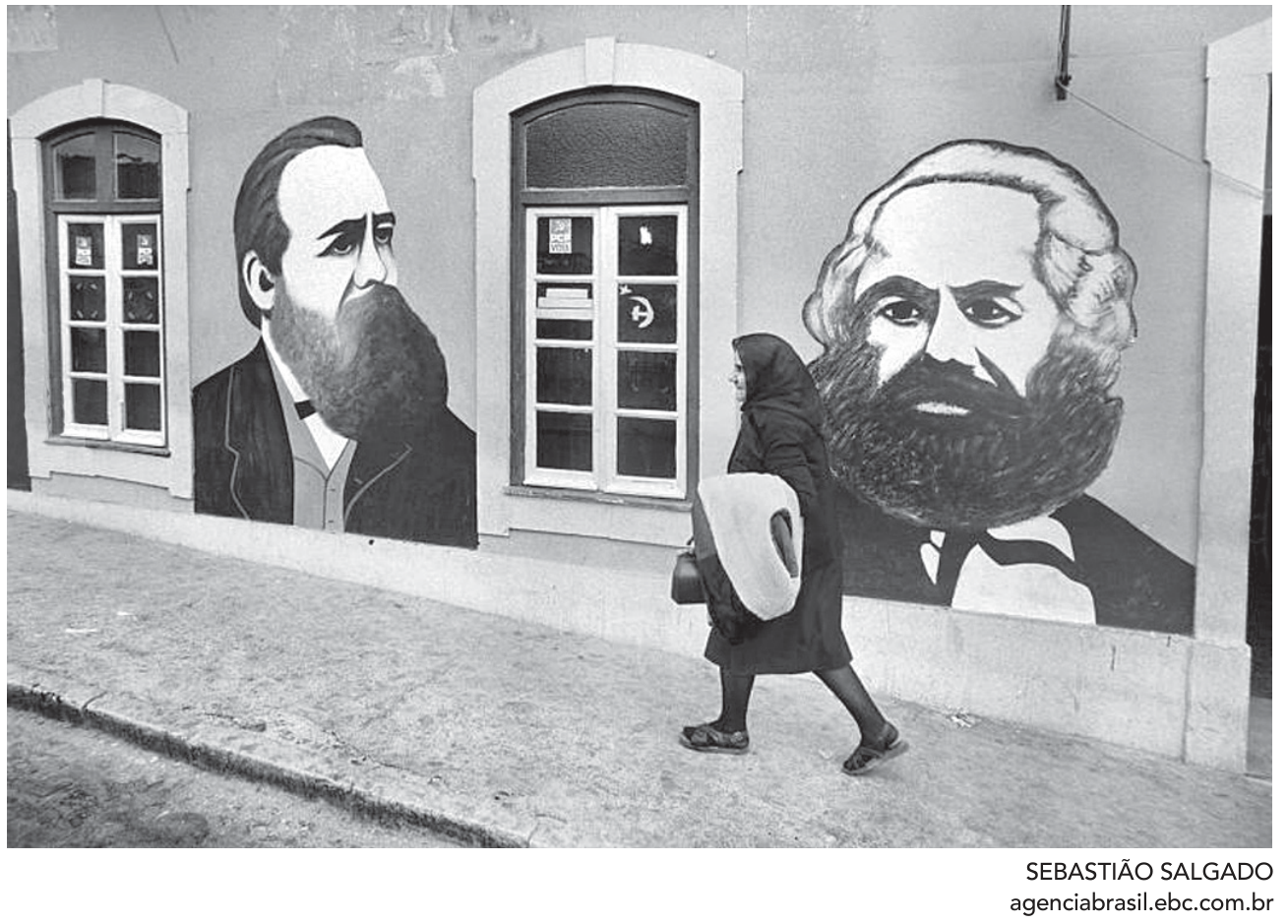 F1 Fotografia em preto e branco de Sebastião Salgado mostrando a fachada de uma casa na vila de Aljustrel com retratos murais pintados de Friedrich Engels e Karl Marx, entre janelas, com cartazes e o símbolo da foice e martelo em uma vidraça, enquanto uma mulher idosa vestida de preto e usando lenço caminha pela calçada.
