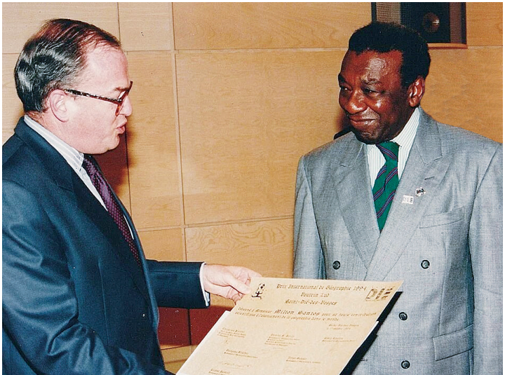 Fotografia de Milton Santos, à direita, de terno cinza, recebendo um diploma entregue por um homem de óculos e terno escuro, à esquerda.