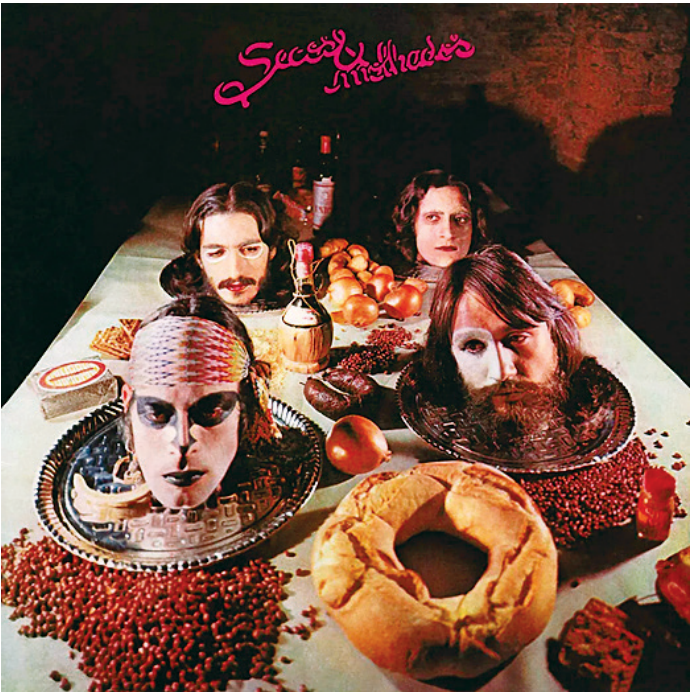 F2 Capa do álbum Secos & Molhados (1973), apresentando uma mesa farta com alimentos e as cabeças dos quatro integrantes maquiados servidas sobre bandejas de prata.