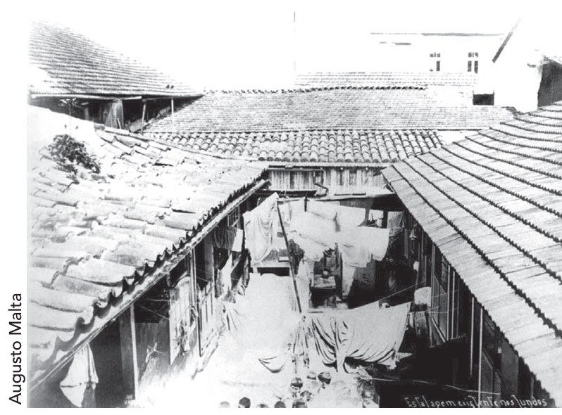 F1 Fotografia em preto e branco de Augusto Malta mostrando a vista aérea de um cortiço com várias construções justapostas, telhados de telha colonial, roupas estendidas em varais no pátio interno e moradores aglomerados.