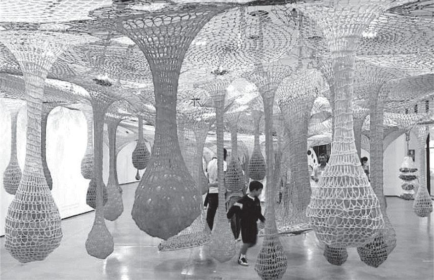 Fotografia da instalação artística Dengo, de Ernesto Neto, exibida no MAM-SP. A sala é tomada por grandes estruturas suspensas de crochê em forma de rede que pendem do teto como estalactites alongadas com bases bulbosas preenchidas. Pessoas, incluindo crianças e adultos, caminham e interagem entre os elementos suspensos da obra.
