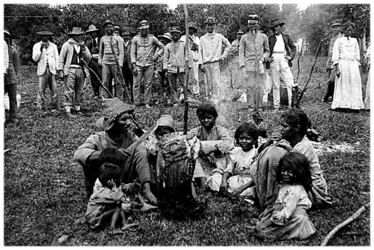 Fotografia histórica em preto e branco de sertanejos e militares na Guerra do Contestado: em primeiro plano, homens, mulheres e crianças sertanejas sentados no chão da mata com roupas desgastadas, enquanto soldados e civis armados permanecem de pé em semicírculo ao redor deles.