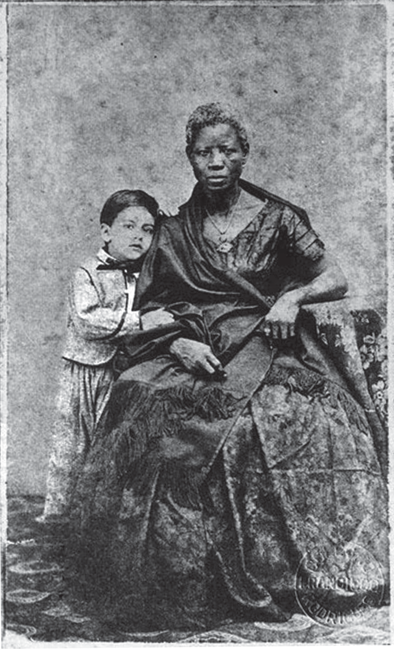 F1 Fotografia em preto e branco de 1860 retratando a ama de leite Mônica, uma mulher negra vestida com trajes formais e joias, sentada em uma poltrona, ao lado do menino branco Augusto Gomes Leal, que está em pé encostado nela.