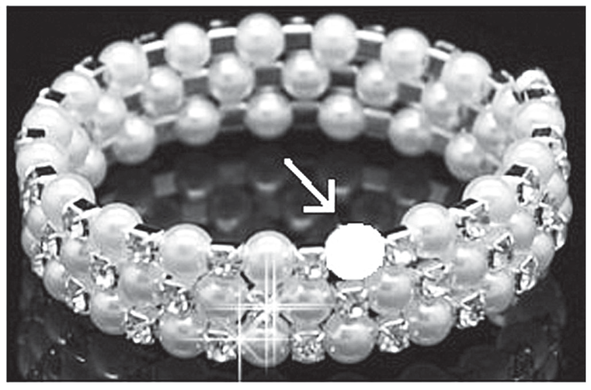 Fotografia de uma pulseira prateada composta por pérolas e pequenos brilhantes dispostos em fileiras; uma seta branca aponta para um círculo branco sólido na fileira superior, indicando a pérola que falta.