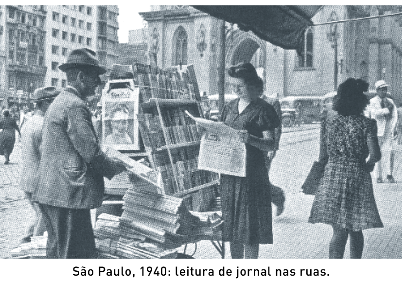 Fotografia em preto e branco de uma banca de jornais em São Paulo, em 1940, com pessoas lendo jornais na rua. Legenda: "São Paulo, 1940: leitura de jornal nas ruas."