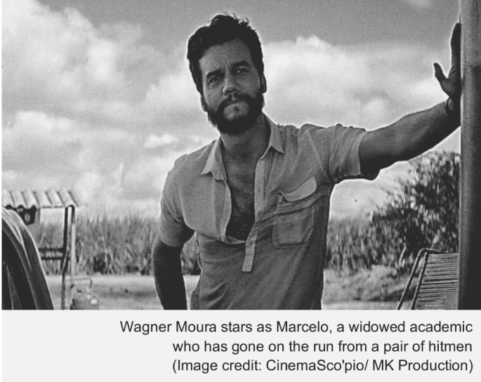 Fotografia em preto e branco do ator Wagner Moura com barba e camisa entreaberta, apoiado com o braço em uma estrutura vertical em uma paisagem aberta. Na parte inferior da imagem, a legenda: "Wagner Moura stars as Marcelo, a widowed academic who has gone on the run from a pair of hitmen (Image credit: CinemaSco'pio/ MK Production)".