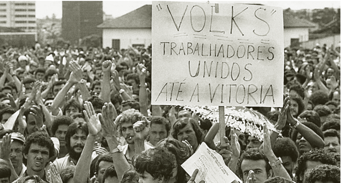 Fotografia em preto e branco de uma assembleia de operários, com uma multidão de braços erguidos. Em primeiro plano, um cartaz com os dizeres: “VOLKS” TRABALHADORES UNIDOS ATÉ A VITORIA.