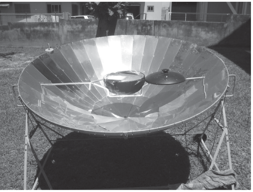 Fotografia de um fogão solar composto por um espelho parabólico côncavo segmentado, montado sobre uma armação de metal, com uma panela suspensa acima de sua região central