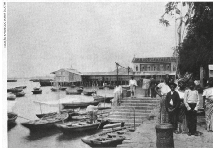 Fotografia em preto e branco retratando o cais de Salvador no início do século XX, com diversas pequenas embarcações na margem e um grupo de pessoas negras em vestimentas da época paradas no cais e na escadaria de acesso à água.