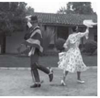 Fotografia em preto e branco de um casal dançando cueca ao ar livre diante de uma casa com telhas coloniais. O homem traja poncho, chapéu e botas com esporas; a mulher usa vestido rodado florido. Ambos seguram um lenço branco erguido durante o passo de dança.