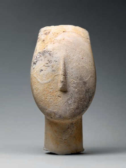 F1 Escultura cicládica de cabeça de figura feminina em mármore, com traços altamente estilizados e minimalistas, destacando-se o relevo do nariz reto e o formato oval e alongado da cabeça sobre um pescoço cilíndrico.