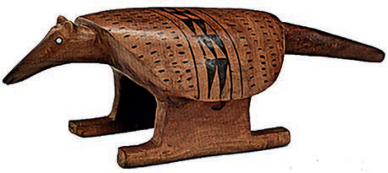 F1 Escultura de banco zoomorfo em madeira no formato de um tatu, com cabeça longa, olhos circulares pequenos, casco arredondado com entalhes pontilhados e grafismos geométricos pintados em preto e vermelho na parte central dorsal, apoiado sobre uma base sólida com pés estendidos e uma cauda cônica comprida.