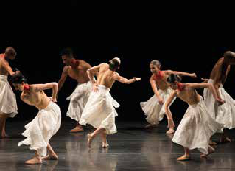 Fotografia do espetáculo de dança Gira, do Grupo Corpo, em que bailarinos e bailarinas com o tronco nu e saias longas brancas dançam e giram pelo palco escuro.