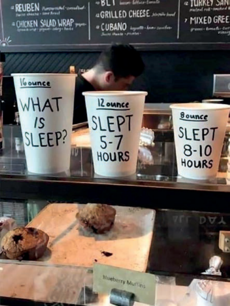 Três copos descartáveis para café de tamanhos diferentes dispostos lado a lado sobre um balcão de cafeteria. O menor copo, à direita, traz o rótulo "8 ounce" e a frase "SLEPT 8-10 HOURS". O copo médio, ao centro, tem o rótulo "12 ounce" e a frase "SLEPT 5-7 HOURS". O maior copo, à esquerda, traz o rótulo "16 ounce" e a pergunta "WHAT IS SLEEP?". Ao fundo, vê-se um funcionário e uma lousa com cardápio.