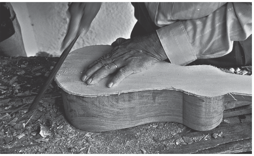 Fotografia em preto e branco mostrando as mãos de um artesão utilizando uma ferramenta manual para lixar ou dar acabamento ao corpo de madeira de um instrumento musical em uma bancada de trabalho coberta de serragem e aparas.