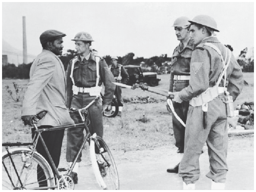 Fotografia em preto e branco de um homem negro ao lado de uma bicicleta sendo abordado por fuzileiros navais armados durante o Apartheid na África do Sul.