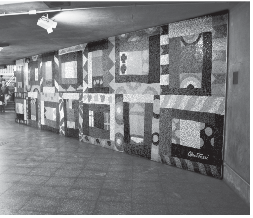 Fotografia de um grande painel de mosaico, intitulado "Colcha de retalhos", instalado na parede de um corredor da estação Sé do metrô. O mosaico é composto por múltiplos painéis retangulares com diversos padrões geométricos e formas estilizadas.