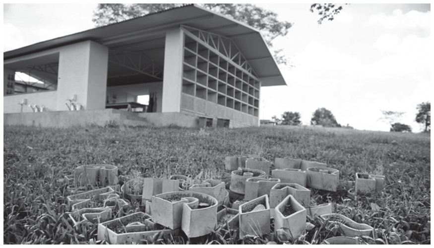 Fotografia da instalação artística em campo aberto, exibindo em primeiro plano diversos vasos de cerâmica com formato de letras espalhados pelo gramado e, ao fundo, um galpão com prateleiras que lembram uma estufa ou ateliê.