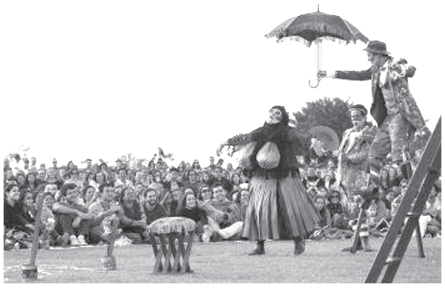 Fotografia em preto e branco de uma apresentação teatral ao ar livre diante de uma plateia sentada no chão. À direita, atores fantasiados atuam, destacando-se um ator sobre pernas de pau segurando um guarda-chuva aberto e outro em trajes bufantes no centro.