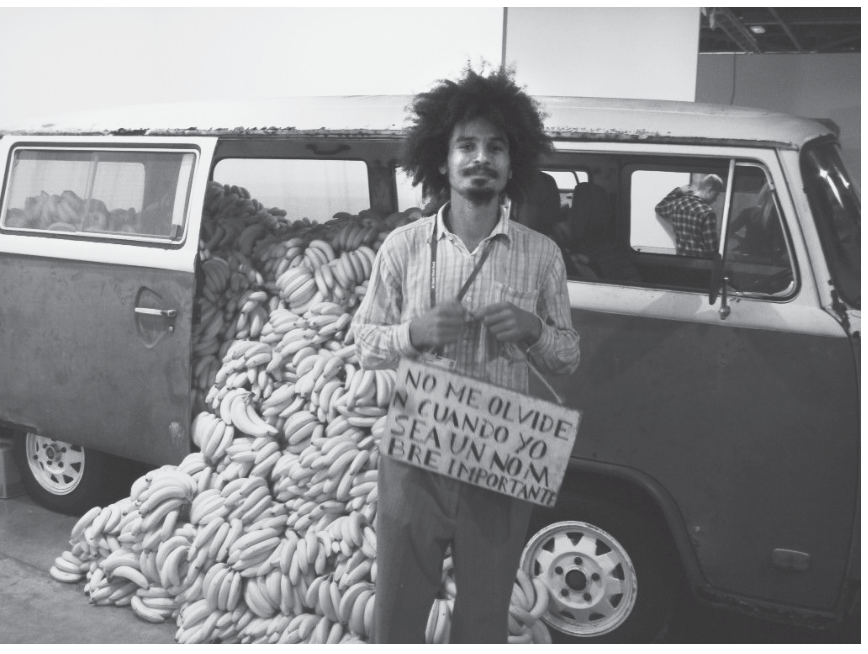 Fotografia em preto e branco do artista Paulo Nazareth em frente a uma Kombi com a porta lateral aberta, de onde transborda uma pilha de bananas até o chão. Ele segura uma placa de papelão no peito onde se lê: "NO ME OLVIDEN CUANDO YO SEA UN NOMBRE IMPORTANTE".