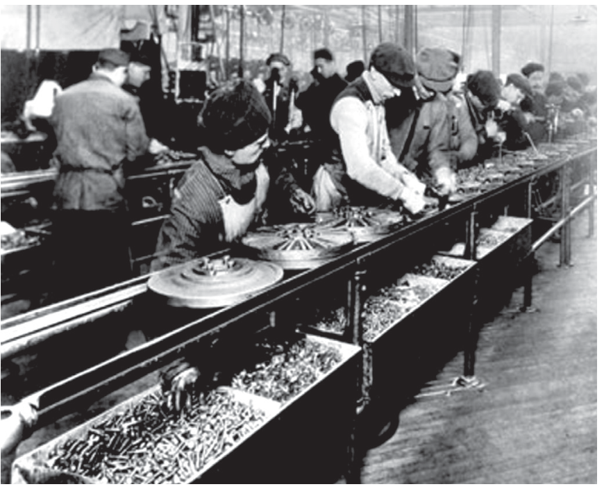 Fotografia em preto e branco do início do século XX mostrando operários trabalhando alinhados ao longo de uma esteira de produção industrial com peças mecânicas e caixas de ferramentas/parafusos.