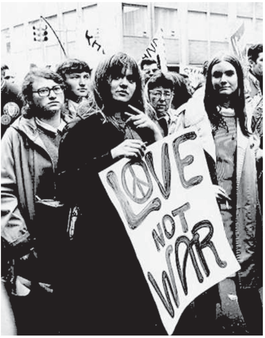Fotografia em preto e branco de um grupo de jovens em protesto na rua; em destaque no centro, uma mulher jovem segura um cartaz onde se lê "LOVE NOT WAR", com o símbolo da paz desenhado no interior da letra "O".