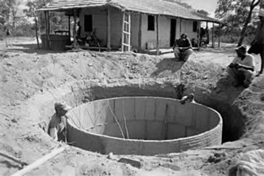 Fotografia em preto e branco mostrando operários em uma área rural construindo uma cisterna cilíndrica de alvenaria em uma grande cava no solo, em frente a uma casa simples.