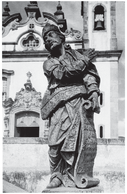 Fotografia em preto e branco de uma das esculturas dos profetas feita por Aleijadinho no Santuário do Bom Jesus de Matosinhos em Congonhas, Minas Gerais, com a igreja e o campanário visíveis ao fundo.