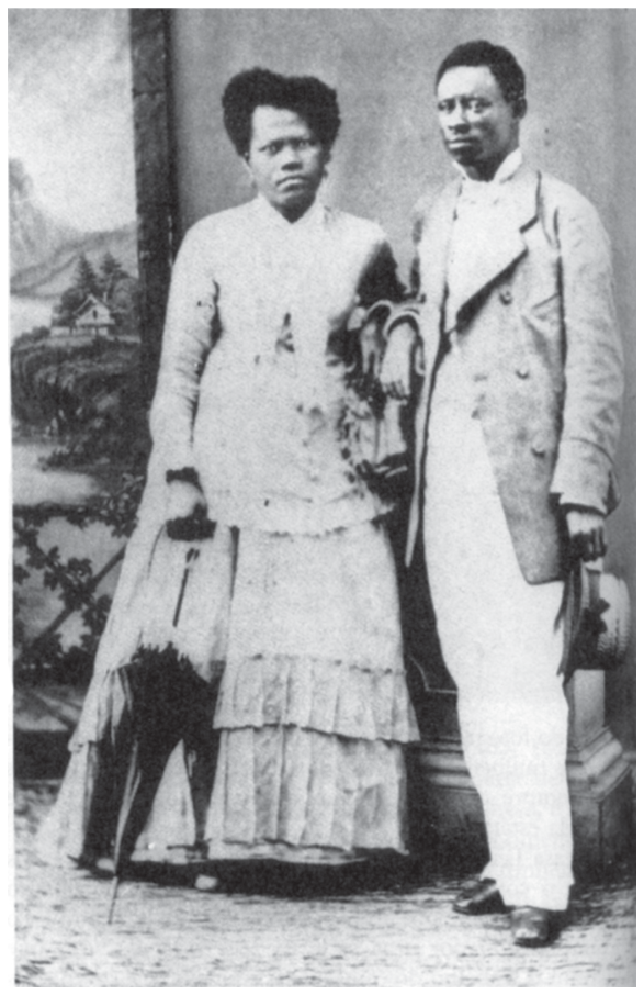 Fotografia em preto e branco de Militão Augusto de Azevedo retratando um casal negro posando em estúdio fotográfico. À esquerda, a mulher veste um vestido longo claro com babados na saia e segura uma sombrinha fechada apoiada no chão. À direita, o homem apoia um dos braços sobre uma mureta ornamental do cenário, veste paletó e calça claros, camisa social, sapatos escuros e segura um chapéu na mão esquerda. O fundo do estúdio exibe uma pintura de paisagem com montanhas e vegetação.