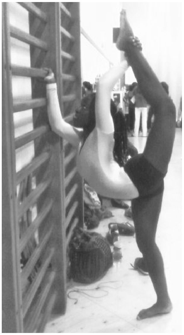 F1 Fotografia em preto e branco de uma ginasta executando um movimento de alongamento extremo: em pé sobre uma perna, ela flexiona a coluna para trás enquanto eleva a outra perna totalmente esticada para cima, segurando um espaldar à sua frente com os braços estendidos.