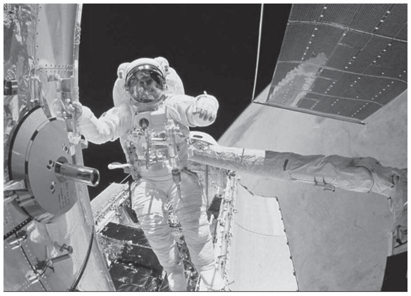 F1 Fotografia em preto e branco de um astronauta com traje espacial realizando manutenção em órbita, próximo a uma estrutura mecânica e aos painéis solares.