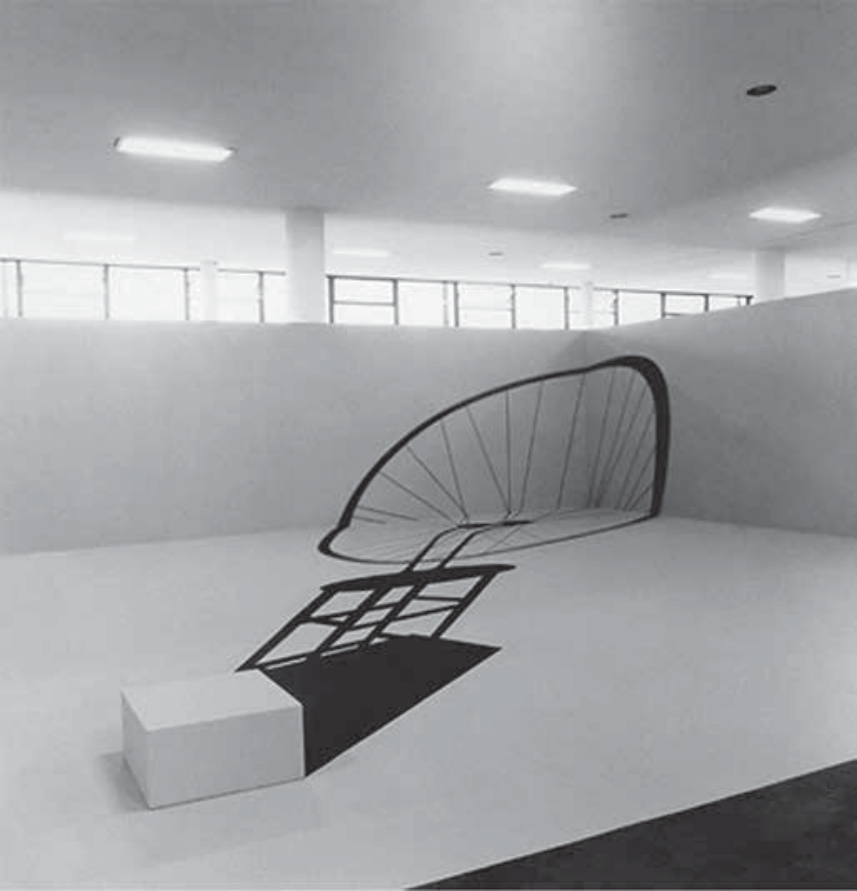 Fotografia da instalação artística In absentia, de Regina Silveira: uma sala branca vazia com a projeção gráfica em perspectiva distorcida de uma sombra representando uma roda de bicicleta sobre um banquinho, que se estende pelo chão e sobe pelo canto da parede; próximo à base da sombra, no chão, há um bloco cúbico branco vazio.