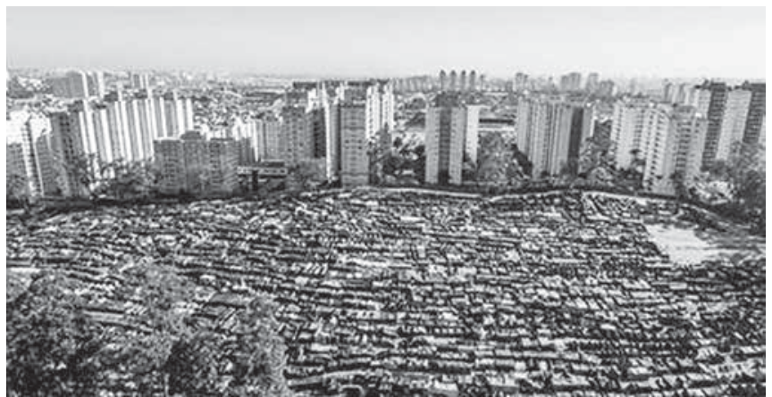 Fotografia aérea de uma vasta área ocupada por inúmeras barracas organizadas lado a lado em um terreno, tendo ao fundo vários edifícios residenciais verticais e a paisagem urbana da cidade.