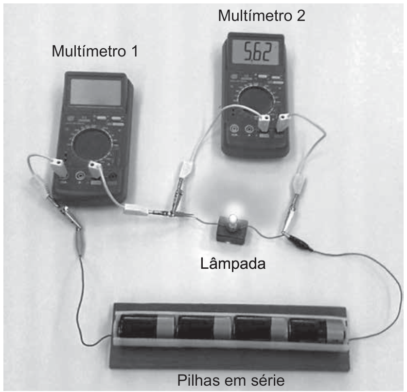 Fotografia de um circuito elétrico montado sobre uma superfície clara. Na parte inferior, há um suporte com quatro pilhas em série. Uma lâmpada de lanterna, que está acesa, fica no centro. O Multímetro 1 está conectado em série no circuito, com a tela apagada (desligado). O Multímetro 2 está conectado em paralelo aos terminais da lâmpada e exibe o número 5,62 no visor digital.