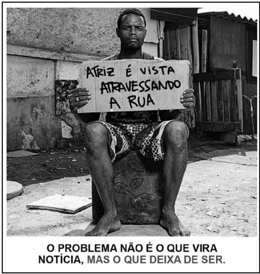 F1 Peça publicitária com a fotografia em preto e branco de um homem sentado em frente a uma habitação precária, segurando um pedaço de papelão com os dizeres: "ATRIZ É VISTA ATRAVESSANDO A RUA". Na parte inferior da imagem, lê-se: "O PROBLEMA NÃO É O QUE VIRA NOTÍCIA, MAS O QUE DEIXA DE SER."