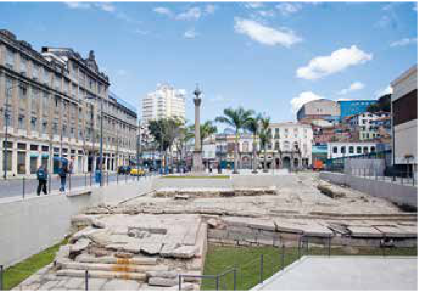 Fotografia do sítio arqueológico do Cais do Valongo, no Rio de Janeiro, com as ruínas de pedra rebaixadas em primeiro plano e, ao fundo, praça com coluna comemorativa, palmeiras e edifícios históricos.