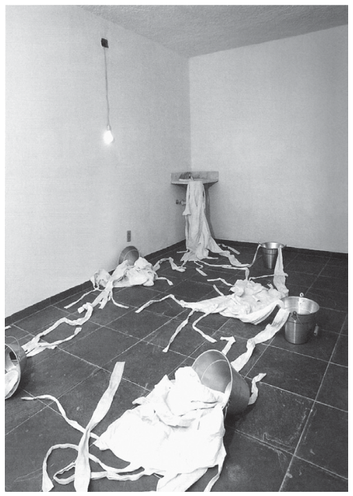 Fotografia em preto e branco da instalação Cry me a river, de Lina Kim. Em uma sala vazia com paredes brancas e piso escuro, vê-se uma pia ao fundo de onde saem camisas de força brancas que se estendem pelo chão até recipientes e baldes de metal espalhados pelo cômodo; uma lâmpada nua pende acesa do teto.